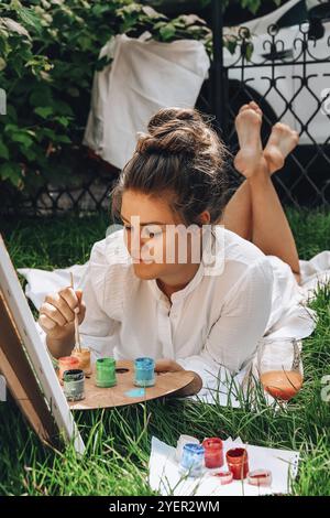 Artiste peint sur le chevalet en plein air dans le jardin. Atelier d'art en plein air. Dessinez sur la toile avec la brosse et la palette en position assise sur la pelouse Banque D'Images