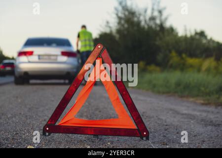 Accident d'urgence d'une voiture de luxe en argent cassé. Conducteur homme installant le panneau d'arrêt triangle rouge sur la route. Automobile de sport allumée des clignotants technique probl Banque D'Images