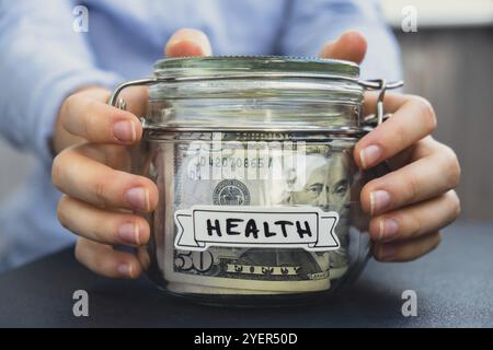 Mains féminines pot en verre plein de billets de banque en dollars américains avec texte SANTÉ. Préparation économisant de l'argent. Consommation et économie modérées. C Banque D'Images
