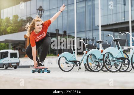 Jeune fille caucasienne blonde s'amusant dehors sur une planche à roulettes, portant des leggings noirs et une chemise rouge, lumière du soleil. Adolescente riant, s'amusant outsid Banque D'Images