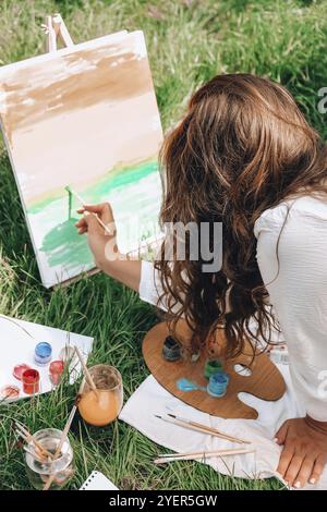 Artiste peint sur le chevalet en plein air dans le jardin. Atelier d'art en plein air. Dessinez sur la toile avec la brosse et la palette en position assise sur la pelouse Banque D'Images