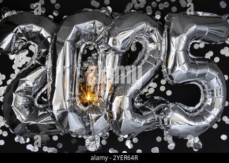 Bonne année 2025 ballons métalliques avec des confettis et des feux d'artifice scintillants Bengale sur fond noir foncé. Carte de voeux argent feuille ballons n Banque D'Images