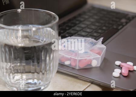 Organisatrice de prises de vue hebdomadaires avec un verre d'eau sur le lieu de travail et ordinateur portable gros plan de la boîte de pilules médicales avec des doses de comprimés pour prendre le médicament quotidiennement Banque D'Images