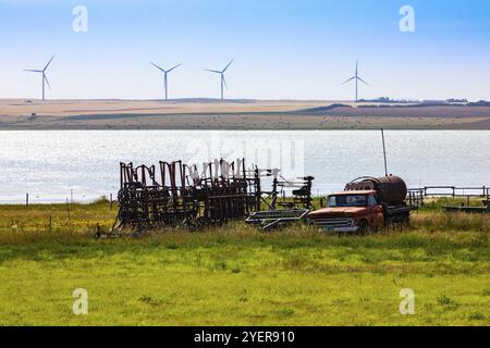 Une vue grand angle de l'équipement agricole rouillé obsolète stocké dans un champ au bord de l'eau. Vieux camions et tracteurs attachés par un lac avec des éoliennes à bac Banque D'Images