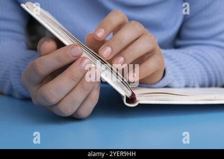 Femme mains manucurées, ongles beiges élégants sur cahier en papier. Gros plan des ongles manucurés de la main féminine en pull bleu sur fond bleu. Hiver ou Banque D'Images