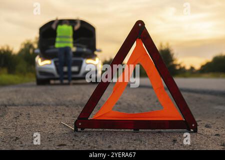 Accident d'urgence d'une voiture de luxe en argent cassé. Conducteur homme installant le panneau d'arrêt triangle rouge sur la route. Automobile de sport allumée des clignotants technique probl Banque D'Images