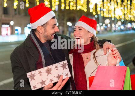 Couple souriant échangeant des cadeaux portant des chapeaux de Père Noël dans une rue festive Banque D'Images