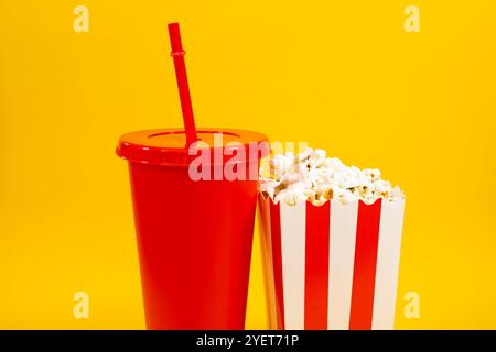 Image horizontale de seau en carton rayé blanc et blanc avec pop-corn et tasse de soude rouge sur fond jaune, concept de nourriture de cinéma et d'entertainm Banque D'Images