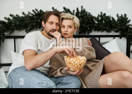 Un couple est assis étroitement ensemble sur leur lit, partageant rire et pop-corn tout en se relaxant. Banque D'Images