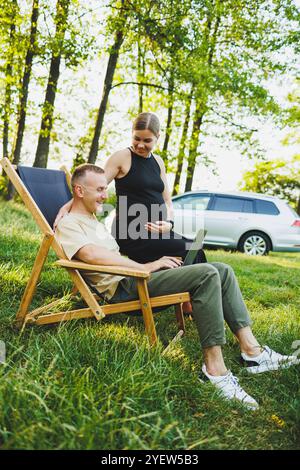 Un homme heureux et sa femme enceinte travaillent sur un ordinateur portable assis dans la nature. Un jeune couple se détache dans la nature et regarde un film sur un lapt Banque D'Images