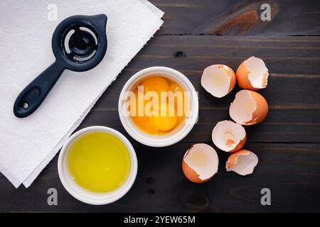 Quatre oeufs séparés dans de petits ramequins blancs : jaunes d'oeufs et blancs d'oeufs montrés avec un séparateur d'oeufs et coquilles d'oeufs sur une table en bois Banque D'Images
