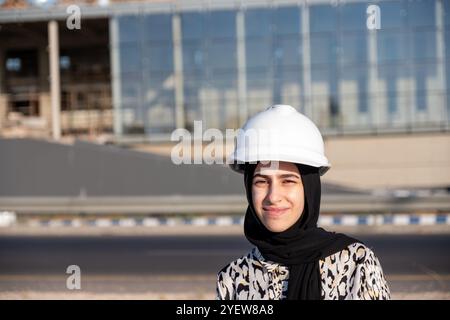 Une ingénieure confiante portant un casque de sécurité se tient sur place, supervisant les travaux de construction. Journée lumineuse et ensoleillée mettant en valeur le leadership et la profession Banque D'Images