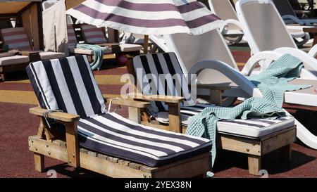 deux chaises longues en bois pour enfants avec couverture en coton doux rayé à côté des chaises longues pour adultes sous parasol en tissu, piscine vide pour la détente Banque D'Images