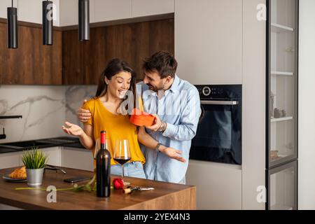 Un couple heureux profite d'un moment cadeau surprise dans une cuisine élégante, entouré de vin et de fleurs, célébrant l'amour et la convivialité. Banque D'Images