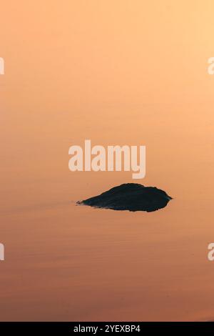 Solitary Rock partiellement submergé dans l'eau calme pendant le coucher du soleil, avec Un gradient d'orange chaud dans le ciel se reflétant sur la surface de l'eau, créant Un Br Banque D'Images