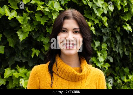 Femme souriante en pull jaune debout à l'extérieur contre des feuilles vertes luxuriantes Banque D'Images