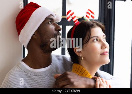 Noël, couple multiracial embrassant par fenêtre, portant des chapeaux festifs, à la maison Banque D'Images