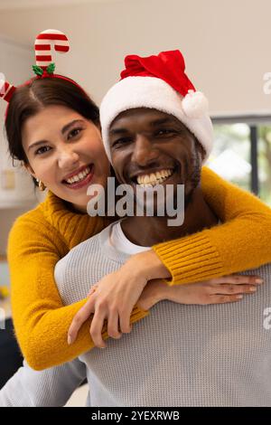 Couple multiracial souriant célébrant Noël à la maison portant des chapeaux et des chandails festifs Banque D'Images