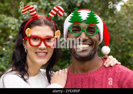 Couple multiracial souriant portant des lunettes et des chapeaux festifs célébrant Noël en plein air Banque D'Images