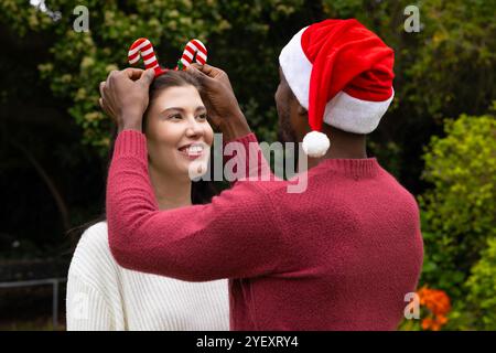 Noël, couple multiracial souriant, portant des chapeaux festifs dans un cadre extérieur Banque D'Images