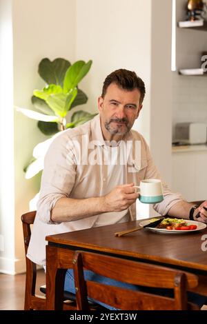homme senior appréciant le petit déjeuner avec du café à la maison, souriant avec satisfaction Banque D'Images