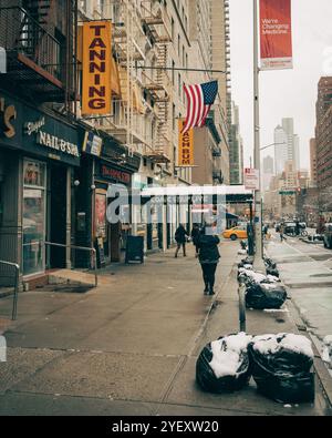 Une scène de rue enneigée dans l'Upper East Side à Manhattan, New York, avec des boutiques, un drapeau américain et des piétons. Banque D'Images