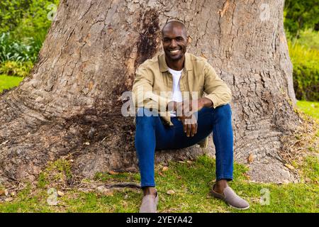 Homme afro-américain souriant assis sous l'arbre, appréciant la nature dans une tenue décontractée Banque D'Images