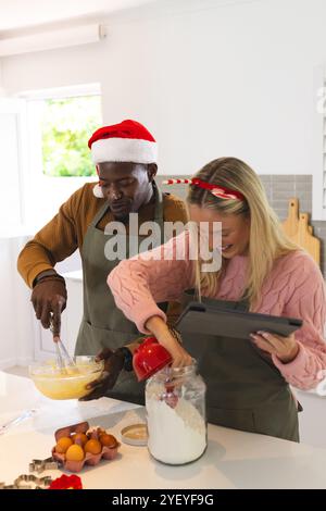 Temps de Noël, cuisson de couple multiracial, portant des chapeaux de vacances festifs, souriant, à la maison Banque D'Images