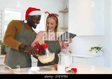 Temps de Noël, couple multiracial faisant des friandises de vacances, souriant et utilisant la tablette, à la maison Banque D'Images