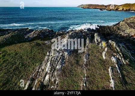 Le littoral accidenté présente des formations rocheuses recouvertes d'herbe verte, tandis que les vagues s'écrasent contre le rivage sous un ciel bleu vif, créant un tranq Banque D'Images