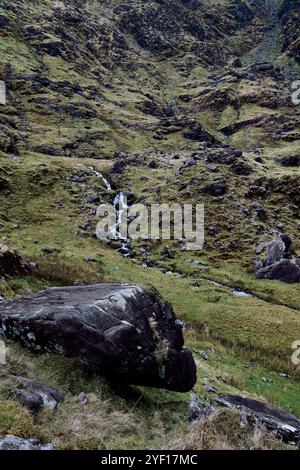 Un ruisseau doux serpente à travers une vallée rocheuse, bordée d'herbe verte vibrante et de rochers éparpillés. Banque D'Images