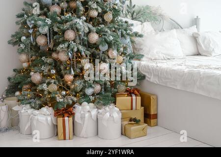 Matin de vacances dans une chambre lumineuse et décorée avec arbre de Noël avec des ornements et des lumières et des boîtes cadeaux emballées rassemblées par le lit avec literie blanche, créa Banque D'Images