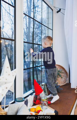 Un garçon de 2 ans est assis sur le rebord de la fenêtre le nouvel an. Ambiance du nouvel an à la maison. L'enfant attend la nouvelle année. Banque D'Images