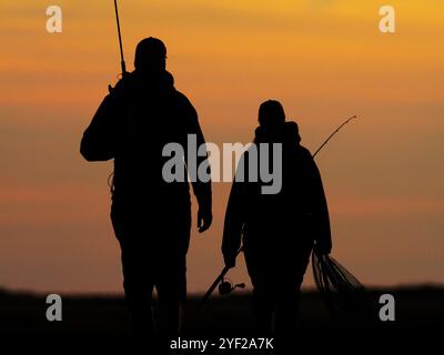 Westermarkelsdorf Auf Fehmarn, Allemagne. 15 octobre 2024. 15.10.2024, Westermarkelsdorf sur l'île de Fehmarn. Un homme et une femme reviennent de la pêche à la truite de mer et marchent sur une digue avec leurs cannes à pêche en contre-jour des derniers rayons du soleil du soir. Ils ne peuvent être vus que comme des silhouettes sombres au crépuscule. Crédit : Wolfram Steinberg/dpa crédit : Wolfram Steinberg/dpa/Alamy Live News Banque D'Images