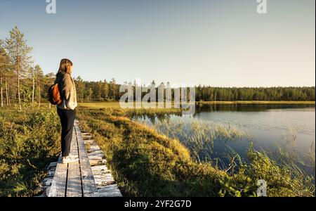 Femme debout sur une promenade en bois, surplombant un lac entouré par la forêt tandis que le soleil du soir jette une lueur chaude sur le paysage de Dalarna Suède Banque D'Images