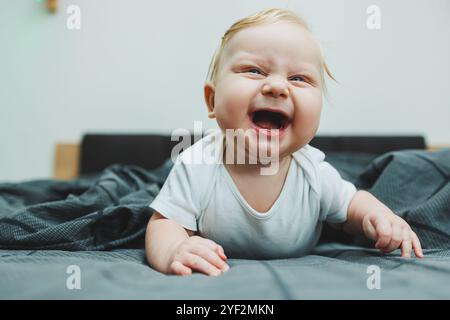 Un bébé garçon joyeux avec de beaux grands yeux est allongé sur le lit à la maison dans un body blanc sur la literie grise. L'enfant est allongé dans son lit à la maison Banque D'Images