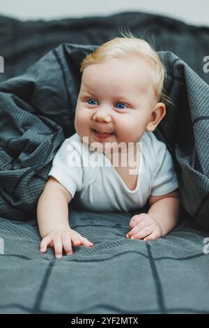 Un bébé garçon joyeux avec de beaux grands yeux est allongé sur le lit à la maison dans un body blanc sur la literie grise. L'enfant est allongé dans son lit à la maison Banque D'Images