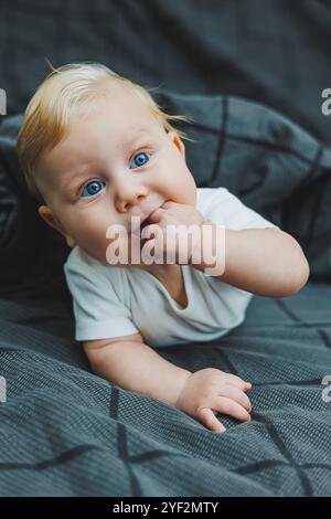 Un bébé garçon joyeux avec de beaux grands yeux est allongé sur le lit à la maison dans un body blanc sur la literie grise. L'enfant est allongé dans son lit à la maison Banque D'Images