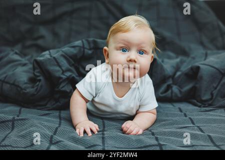 Un bébé garçon joyeux avec de beaux grands yeux est allongé sur le lit à la maison dans un body blanc sur la literie grise. L'enfant est allongé dans son lit à la maison Banque D'Images