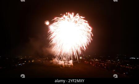 Un grand feu d'artifice blanc explose dans un ciel nocturne sombre. Il y a une petite ville ou une ville au loin avec des lumières éclairant le paysage. Banque D'Images