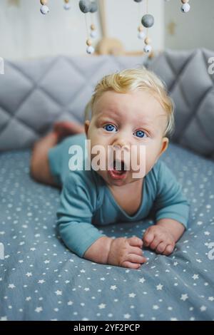 Un enfant de 6 mois est allongé dans un berceau et regarde la caméra. Linge de lit gris pour un lit bébé Banque D'Images