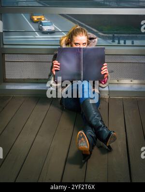 Une femme est assise sur un plancher en bois, se cachant partiellement derrière un grand livre ouvert. Une fenêtre derrière révèle une vue d'une autoroute avec des véhicules qui passent. Banque D'Images