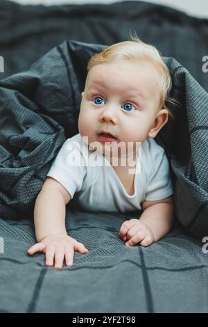 Un bébé garçon joyeux avec de beaux grands yeux est allongé sur le lit à la maison dans un body blanc sur la literie grise. L'enfant est allongé dans son lit à la maison Banque D'Images