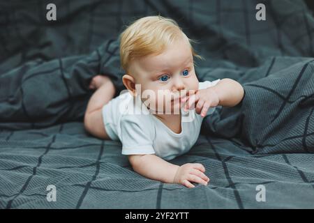 Un bébé garçon joyeux avec de beaux grands yeux est allongé sur le lit à la maison dans un body blanc sur la literie grise. L'enfant est allongé dans son lit à la maison Banque D'Images