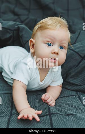 Un bébé garçon joyeux avec de beaux grands yeux est allongé sur le lit à la maison dans un body blanc sur la literie grise. L'enfant est allongé dans son lit à la maison Banque D'Images