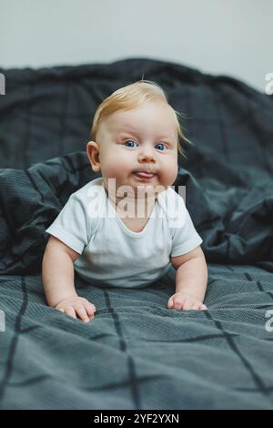 Un bébé garçon joyeux avec de beaux grands yeux est allongé sur le lit à la maison dans un body blanc sur la literie grise. L'enfant est allongé dans son lit à la maison Banque D'Images