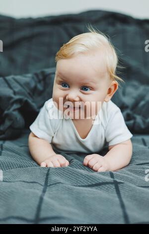 Un bébé garçon joyeux avec de beaux grands yeux est allongé sur le lit à la maison dans un body blanc sur la literie grise. L'enfant est allongé dans son lit à la maison Banque D'Images