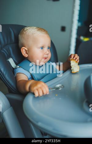 Un garçon de 1 an est assis dans une chaise haute et mange du pain. Le bébé mange du pain. L'auto-alimentation de l'enfant. Banque D'Images