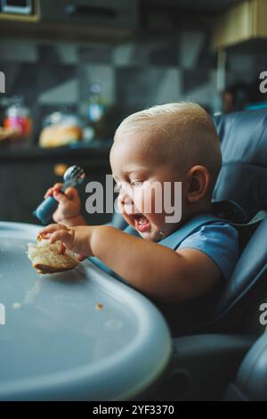 Un garçon de 1 an est assis dans une chaise haute et mange du pain. Le bébé mange du pain. L'auto-alimentation de l'enfant. Banque D'Images