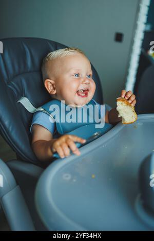 Un garçon de 1 an est assis dans une chaise haute et mange du pain. Le bébé mange du pain. L'auto-alimentation de l'enfant. Banque D'Images